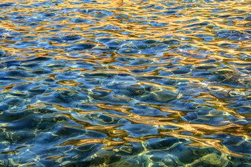 Background with abstract patterns on the water surface. Evening sun plays on the water, blue mixes with yellow-orange tones and forms abstract structure.