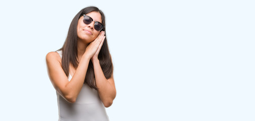 Young beautiful hispanic wearing sunglasses sleeping tired dreaming and posing with hands together while smiling with closed eyes.