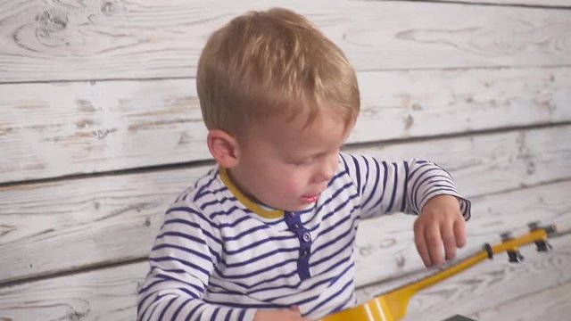 happy one year old boy plays his guitar or ukulele and sings songs, sitting in the wooden room.