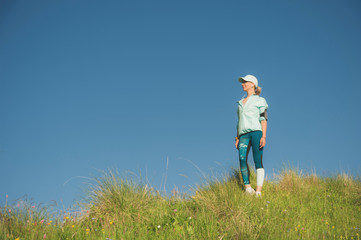 Naklejka na ściany i meble Fitness runner woman listening to music on the nature. Portrait of beautiful girl wearing earphones earbuds and running cap. against the background of a green field and blue sky