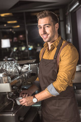 handsome young barista preparing coffee with coffee machine in cafe and looking at camera