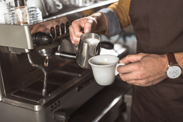 cropped shot of barista in apron pouring milk into coffee while preparing it in cafe