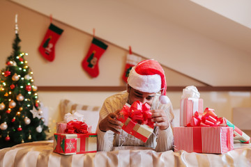 Fototapeta premium Mixed race man with santa's hat on head lying on bed and trying to open Christmas gift. Around him presents, in background Christmas decorations.