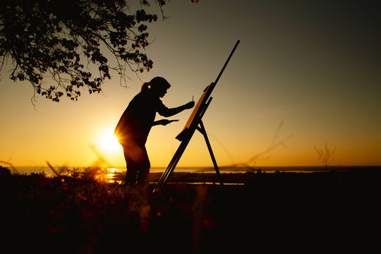 silhouette of a woman painting a picture with paints on canvas on an easel, girl with paint brush and palette engaged in art on the nature in a field at sunset