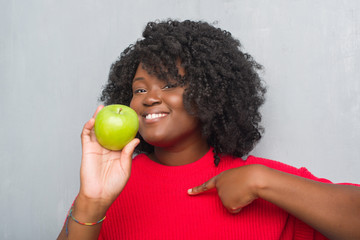 Young african american woman over grey grunge wall eating green apple with surprise face pointing finger to himself