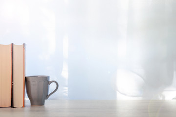 A wooden desk with thick books and a gray ceramic mug with coffee set under the window. Copy space
