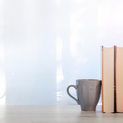 Real minimal photo of a wooden desk with thick books and a gray ceramic mug with coffee set under the window. Copy space