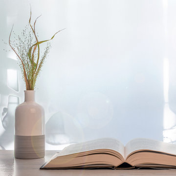 Wooden desk with open thick book and a white vase with green grass set under the window. Copy space