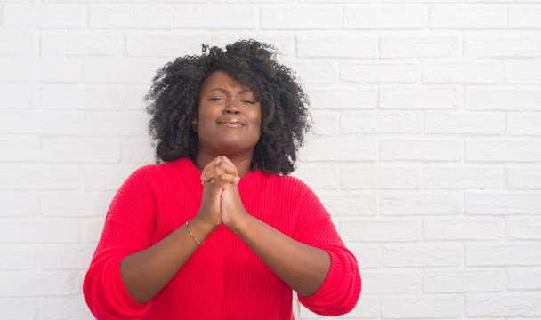 Young african american plus size woman over white brick wall begging and praying with hands together with hope expression on face very emotional and worried. Asking for forgiveness. Religion concept.