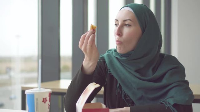 Young muslim woman in hijab sitting at the table by the window in fast food and eating potatoes