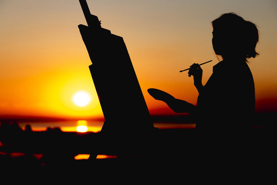 silhouette of a woman painting a picture on canvas on an easel, girl with paint brush and palette engaged in art in a field at sunset