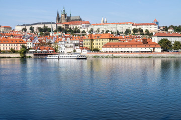 Obraz premium Prague castle and old town cityscape with vltava river and tour boat on a sunny day, a tour boat navigates the calm waters, reflecting the surrounding buildings and vibrant red rooftops