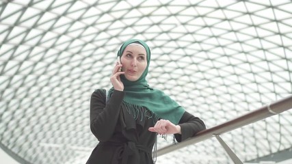 Pretty muslim woman in a hijab with a backpack is standing in a shopping center and talking on the phone,slow mo
