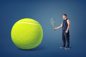 An athletic man stands in a side view holding a tennis racquet in front of a giant yellow ball.