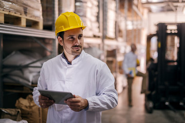 Picture of young focused male manager standing in factory and holding tablet. Dressed in white coat with helmet on his head. Looking satisfied and proud.