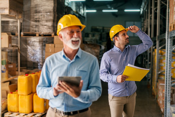 Picture of father and son walking through their warehouse with helmets on their heads. Son in looking and big shelf full of products and looking focused.