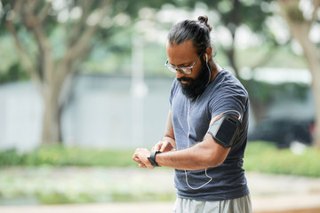 Bearded athletic Indian man in glasses checking time on smartwatch while having workout outdoors on...