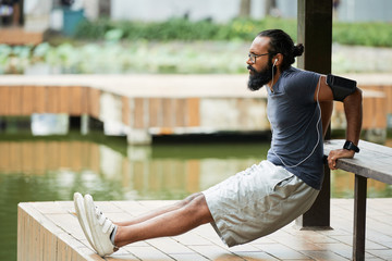 Fototapeta na wymiar Side view of determined bearded Indian man doing warming up exercise outdoors and listening to music with earbuds