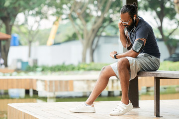 Bearded Indian sportsman looking at watch and taking his pulse after running while sitting on bench outdoors