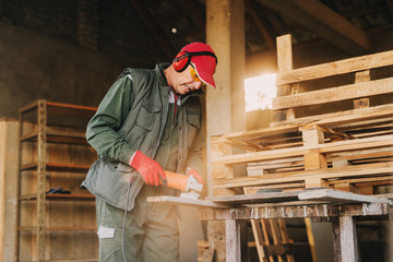 Picture of mature carpenter man in protective uniform shaping wood with electric grinder. Enjoying his job in his working garage on sunny day.