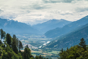 Fototapeta na wymiar Rhine Valley in Switzerland. View of the river and residential houses in the valley. The sky is covered with clouds. Alps are on the right in the picture and in the background.