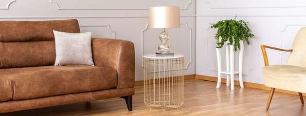 Panoramic view of stylish lamp on small elegant table next to brown leather couch with pillow in...
