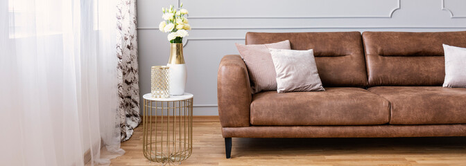 Panoramic view of white roses in elegant vase standing on the stylish table with marble countertop next to big brown leather sofa with pillows, real photo