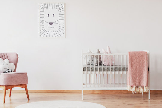 White crib with pillows and pink blanket in cute baby room with posters on the wall and pink armchair with cloud shape pillow