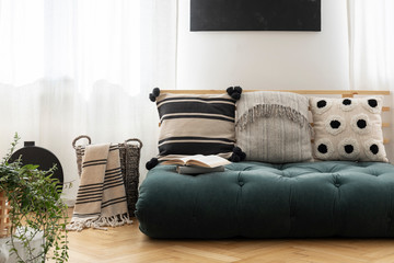 Patterned cushions on green futon in bright living room interior with blanket in the basket. Real photo