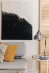 Grey lamp on table next to sofa with pillows in modern living room interior with poster. Real photo