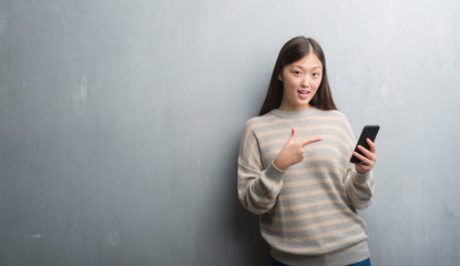 Young Chinese woman over grey wall looking at smartphone very happy pointing with hand and finger