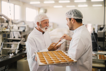 Picture of two employees in sterile clothes in food factory smiling and talking. Younger man is holding tray full of fresh cookies while the older is holding tablet and checking production process.