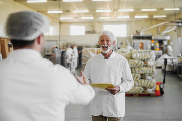 Obraz na płótnie Canvas Picture of mature cheerful older man in sterile clothes standing in food factory and being happy about seeing his colleague. Team work and good job concept.