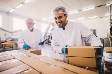 Two male colleagues i sterile clothes preparing boxes with products for transport. Standing in bright room or warehouse and counting package. - obrazy, fototapety, plakaty