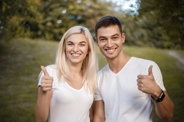 Happy young cute loving couple standing on grass in nature green park with beautiful sundown light. Looking camera showing thumbs up gesture.