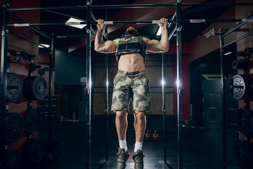 Muscular caucasian bearded man doing pull-ups in military style weighted vest in crossfit gym....