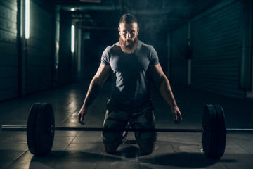 Fototapeta na wymiar Muscular attractive caucasian bearded man in corridor looking at camera, on knees, ready to lift barbell.