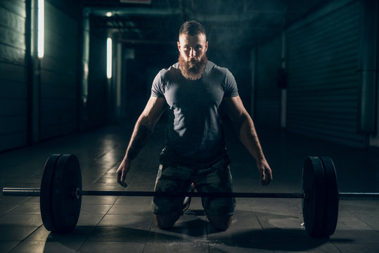 Muscular attractive  caucasian bearded man in corridor looking at camera, on knees, ready to lift barbell.
