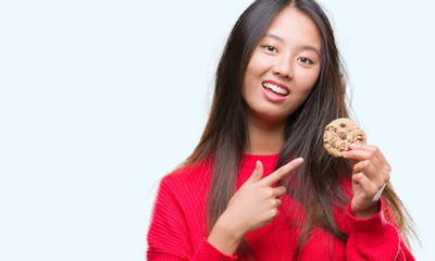Young asian woman eating chocolate chip cookie over isolated background very happy pointing with hand and finger
