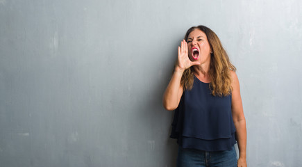 Middle age hispanic woman standing over grey grunge wall shouting and screaming loud to side with hand on mouth. Communication concept.