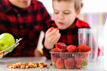 Young father and cute son sitting by the kitchen table full of fresh fruits. Boy is eating strawberries.