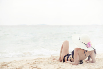 tanning suntanned young woman in bikini wearing straw hats lying on a tropical beach on white sand stretching up slender leg Blue sea in the background. Summer holiday fashion vacation travel concept.