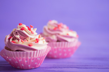 Closeup two cupcakes with creamy pink and white top decorated with little hearts on purple wooden background.