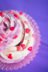 Closeup cupcake with creamy pink and white top decorated with little hearts on purple background.