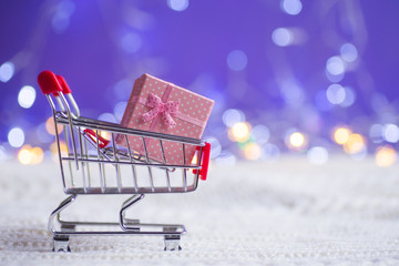 Pink gift box in small supermarket trolley on white knitted fabric on purple background with warm bokeh.