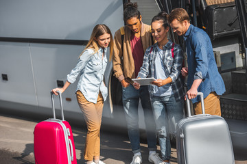 asian young woman showing digital tablet to multicultural friends with wheeled bags near travel bus at city street