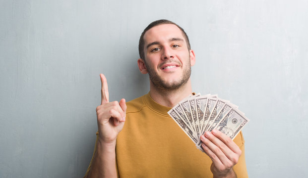 Young caucasian man over grey grunge wall holding dollars surprised with an idea or question pointing finger with happy face, number one