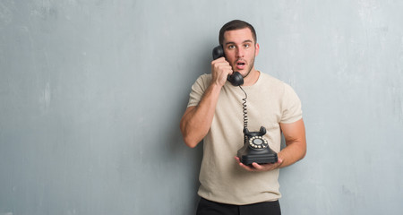 Young caucasian man over grey grunge wall calling using vintage telephone scared in shock with a surprise face, afraid and excited with fear expression