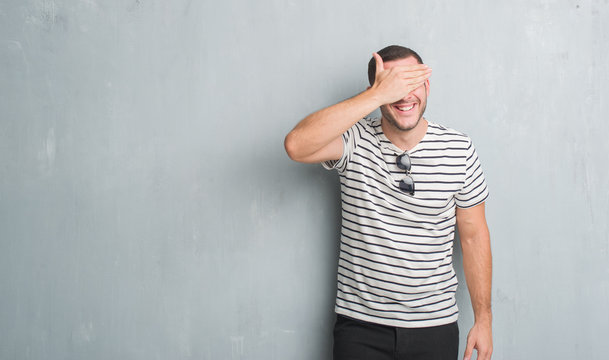 Young caucasian man over grey grunge wall smiling and laughing with hand on face covering eyes for surprise. Blind concept.