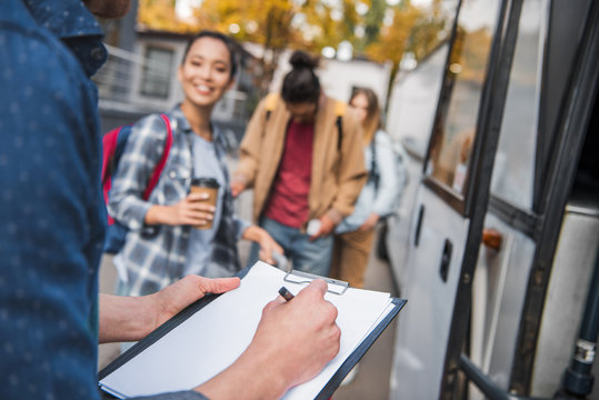 cropped image of travel bus controller writing in clipboard while tourists standing near bus at city street